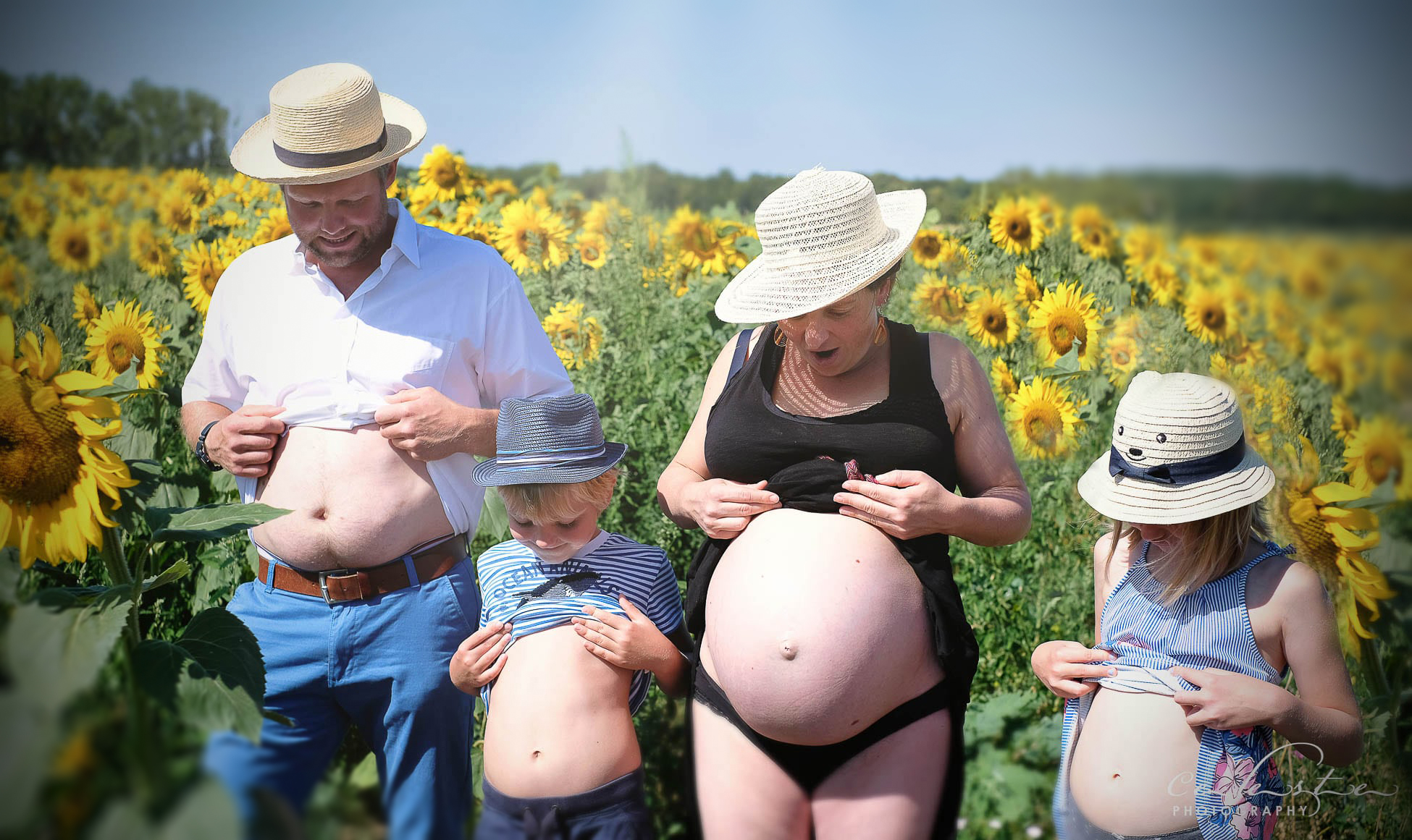 un couple avec leurs enfants dans un champs de tournesol regardent tous leurs ventres gonflés d'un air surpris (la mère est enceinte)